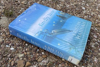 Library book, Firefly Lane by Kristin Hannah, laying on gravel.