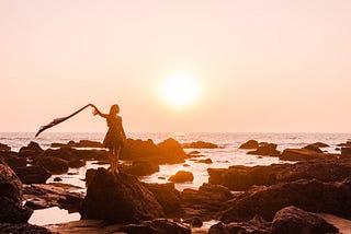 A woman stands on a rock-strewn beach as the sun sets over the ocean.