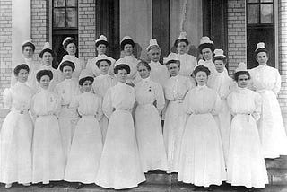 20 women in white nursing uniforms stand outside of Naval Hospital building, 1908.