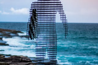 Metal, mesh-like sculpture of surfer standing with board overlooking the ocean.