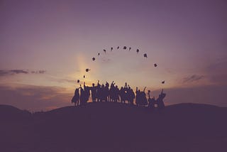 Group of silhouetted graduates in their gowns, standing on top of a hill at dawn, caps thrown up in the air forming a celebratory rainbow.