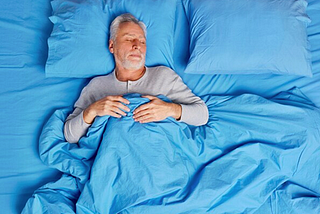 A photo of an older man asleep in a bed with blue sheets, pillowcases, and covers.