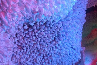 This is a multi-colored photo of a large art installation made from recycled trash bags. The piece is made to look like a coral reef. The trash bags are white but are surrounded by blue, pink and purple lighting.