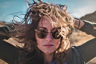 girl with brown hair wearing sunglasses and dark top with hands behind her head