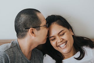 Image of couple sitting next to each other. She’s smiling and looking down while leaning towards him. He’s in profile, kissing her forehead.