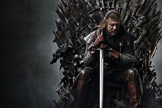 Eddard Stark sits on the Iron Throne. He gazes down. Both hands grip his sword, its point planted in the ground.