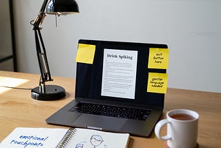 A laptop with text about drink spiking, next to a lamp on a desk. There are yellow sticky notes on the screen with ‘exit button here’ and ‘gentler language needed’. A notebook reads ‘emotional touchpoints’. On the right is a white mug with tea or coffee. This suggests the desk of a trauma-informed content specialist.
