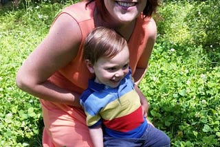 Smiling woman in coral dress has a smiling toddler on her lap, kneeling in a bed of clover.