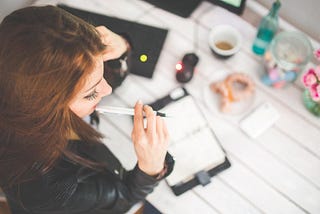 Young woman at her desk, looking at her diary while holding a pen. Photo taken from above.