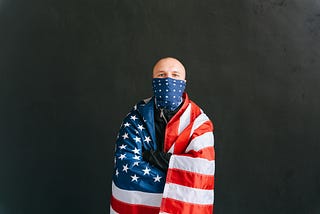 A bald Caucasian male stands center with his arms crossed. An American flag is wrapped around his face (like a bandana) and his entire body.