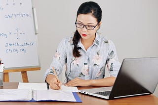 A woman sitting a desk writing on paper with a pencil while listening through headphones to something on her laptop