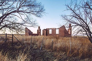 The crumbling brick remains of a building with two window outlines, two visible chimneys, and a missing roof framed between two small barren trees behind a barbed wire fence and in the middle of waist-high dead grass