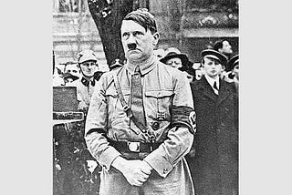 Hitler is standing in his uniform, hands folded.