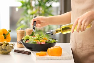 A green salad with olive oil being poured on it by a woman. Pumpkin, olives, pepper mill and peppers are in the background.