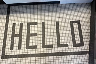 The image shows a tiled floor with the word “HELLO” spelled out in large, black, capital letters. The tiles are small and square, with a white background and black lines forming the letters and a border around the word. The word “HELLO” is centered within the border.