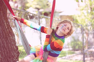 Image of a child on a swing, wearing a rainbow shirt, tutu and rainbow make-up