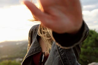 A person with long hair puts their hand out toward the viewer, blocking the view of their face.