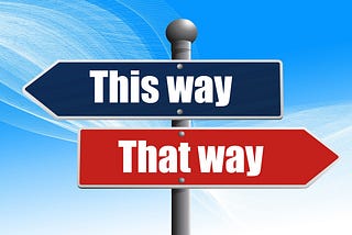 This is metal sign post with two directional arrows on it. On top is a blue one with the words, “This way” (pointing to the left), and below it is a red one with the words, “That way” (pointing toward the right). The background is sky blue with swishes coming down diagonally from top left to bottom right.