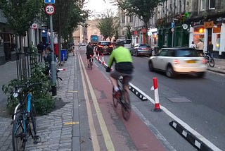 Image shows Forrest Rd in Edinburgh, a narrow one-way street near to the city centre. There are shops & cafes on both sides and the left hand part of the carriageway is coloured red and protected by plastic bollard (or wands) to create a segregated cycle lane. There are two cyclists riding in the bike lane, and several cars driving in the main carriageway. The street is busy with parked vehicles, buses, cars, pedestrians and cyclists.