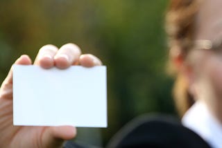 A blank business card is the main focus of the image, being held out by a lady who is fuzzily in the background.