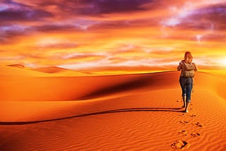 woman hikes across dessert, which is in orange tones with a pink and purple sky