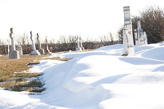 photograph of an old cemetary in winter, featuring drifts of snow against a stark sky