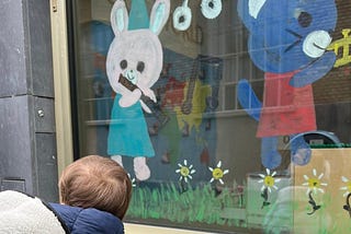 A kid in his troller is in front of the window of his kindergarten, where a bunny and a rat are painted