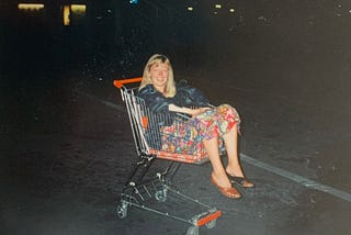 A laughing woman dressed in a black leather jacket and colourful dress lying in a supermarket shopping trolley, with her legs dangling out over the front.