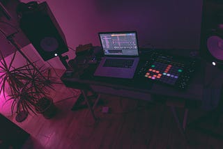 Foto de uma mesa com equipamentos de produção musical e um notebook. Ao redor, na sala, estão caixas de som e uma planta.