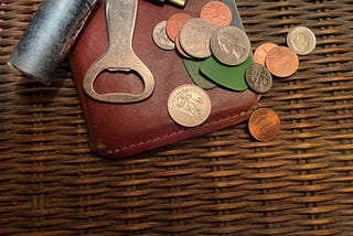 A collection of personal items on a wicker end table, including a set of keys, a tan leather pocket notebook holder, two green guitar picks, and a handful of coins, with a single quarter set aside, ready for decision-making duty.