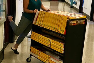 A woman with short dark hair pushes a cart of 100 World History textbooks with yellow spines. She is using her full weight, but posing for the camera with one foot kicked up.
