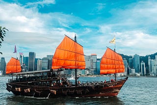 A junk rig, a traditional Chinese ship, sailing across the river connecting Hong Kong’s island from the mainland. The ship has bright orange sails that stand out from the partially cloud sky.