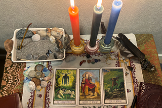 Altar with sand, rocks and seashells from Norwegian beach, currency from around the world, ayahuasca vine, shells and other stones from across the globe, candles, feathers and crystals to represent all chakras, abundance, grace and freedom. The cards are from the Druidcraft deck. XV Cerunnos, Queen of Wands and Ten of Wands.