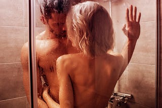 A sexy young woman with platinum blonde hair and a well-built, dark haired man with a beard embrace in a shower.