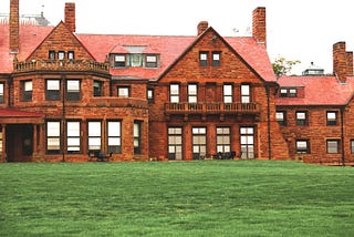 Large estate building with 4 chimneys and 20 windows visible on the two stories. Constructed of brick with two balconies and a large green lawn.