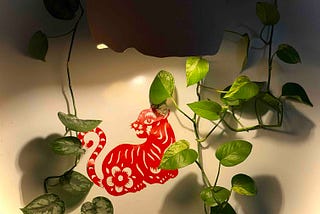 A red paper‑cut cat with floral patterns hangs on a light wall, surrounded by trailing green pothos vines that frame the artwork with soft, natural curves.