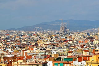 A colorful city spread out with low rise buildings — in the distance a brown cathedral under construction towers over the city. A hazy mountain is visible in the background.