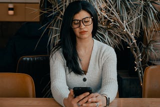 A woman with dark hair and glasses sits in an office boardroom and is distracted by her phone.