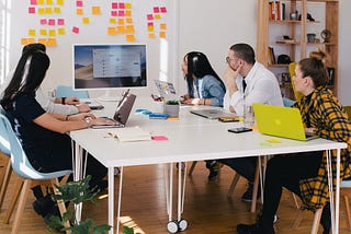 A team of 5 people sitting around a table and watching a presentation on a monitor.