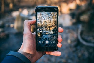 A hand holding an iPhone that is in photo-taking mode with an image of the sun-lit river and forest behind the iPhone.