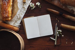 In the middle of a wood table there is a notebook wide open, the pages of it are blank. In the left there are some pens and pencils, and in the right corner there’s something that looks like a croissant.