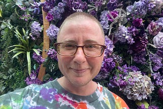 The author is standing in front of a wall of purple flowers and leaves. His hair is cut very short, and he is wearing brown glasses. He is smiling slightly, but it looks a little forced. He is wearing a colorful tie dye shirt.