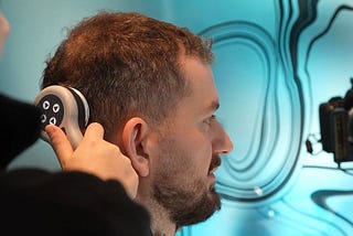 A clinician uses a handheld trichoscopy device to examine the hair above a man’s ear, the man shown in profile in a clinic.
