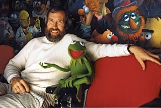 Uma fotografia do Jim Henson (criador dos Muppets) sentado num sofá vermelho junto com o sapo Caco (Kermit).