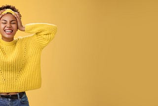 Carefree, happy, attractive girl in a yellow sweater and headband, touching head and smiling broadly. Yellow background.