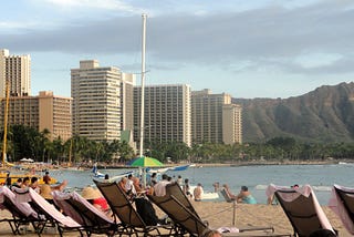 Lounge chairs on the beach with a sailboat (without a sail) in the water. There are not a lot of people on the beach, and it appears to be late afternoon. There are tall buildings (hotels) and a mountain (Diamond Head) in the background. The beach is bordered by lush palm trees.