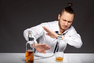A man in a dress shirt and time sitting at a dinner table shelters himself from the glass and bottle of scotch in front of him that is shackled to his wrist