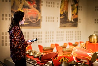 A visitor, side profile, wearing headphones and holding a smartphone, is focused on an audio guide. They stand before a vibrant exhibit of traditional red and gold musical instruments, with a large mural in the soft-focused background.