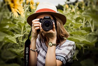 Vrouw fotograaf fotografeert met een digitale Canon camera en kijkt richting de lens. Er hangt een camera strap met EOS en ze staat in een veld met zonnebloemen. De beginnend fotograaf kijkt door de lens om een foto te maken en stelt schep door de lens te draaien. Deze foto hoort bij het artikel ‘welke digitale camera is het beste voor beginners?’ om een keus te maken bij het kiezen van een camera.