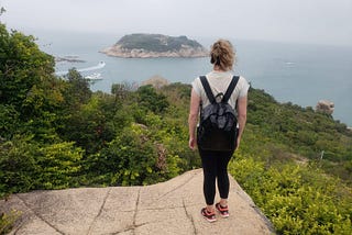 Alice stands on the edge of a cliff overlooking greenery and the ocean.